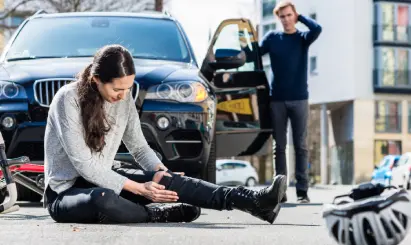 Injured woman sitting on street after bicycle accident with car; driver in background appears distressed.